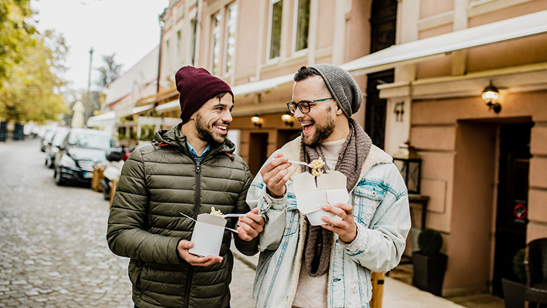 Two men walking down a cobbled street, laughing and eating out of takeout containers