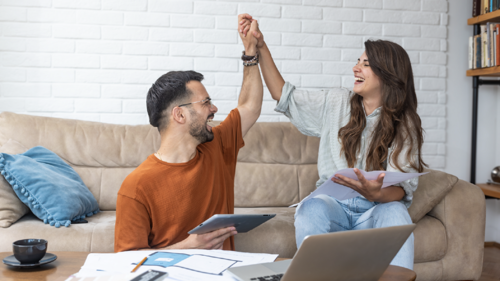 Couple sit on couch high-fiving in front of laptop and papers celebrating affordable payments